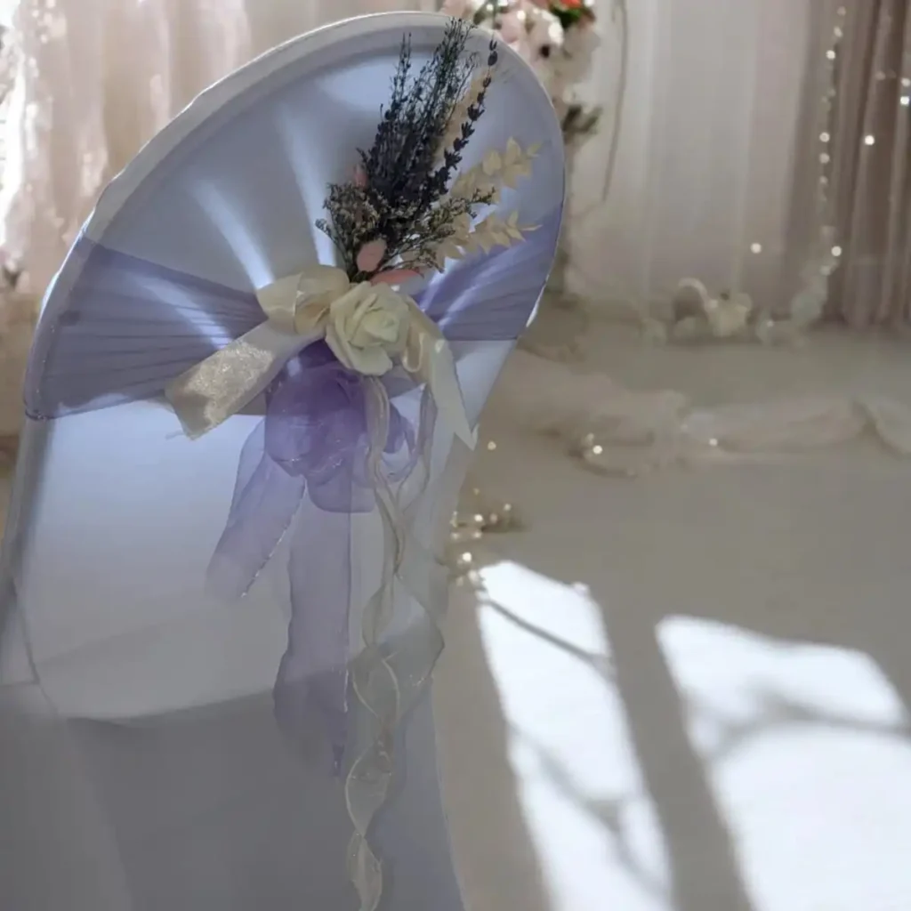 Décoration florale pour chaise de cérémonie laïque, composée de rubans en organza violet et ivoire, ornée de fleurs artificielles délicates. La chaise est recouverte d'une housse blanche, créant une ambiance élégante et raffinée. En arrière-plan, des rideaux et des lumières douces ajoutent à l'atmosphère romantique de l'événement. Parfait pour une allée de mariage, cette décoration apporte une touche sophistiquée et harmonieuse à la cérémonie.
