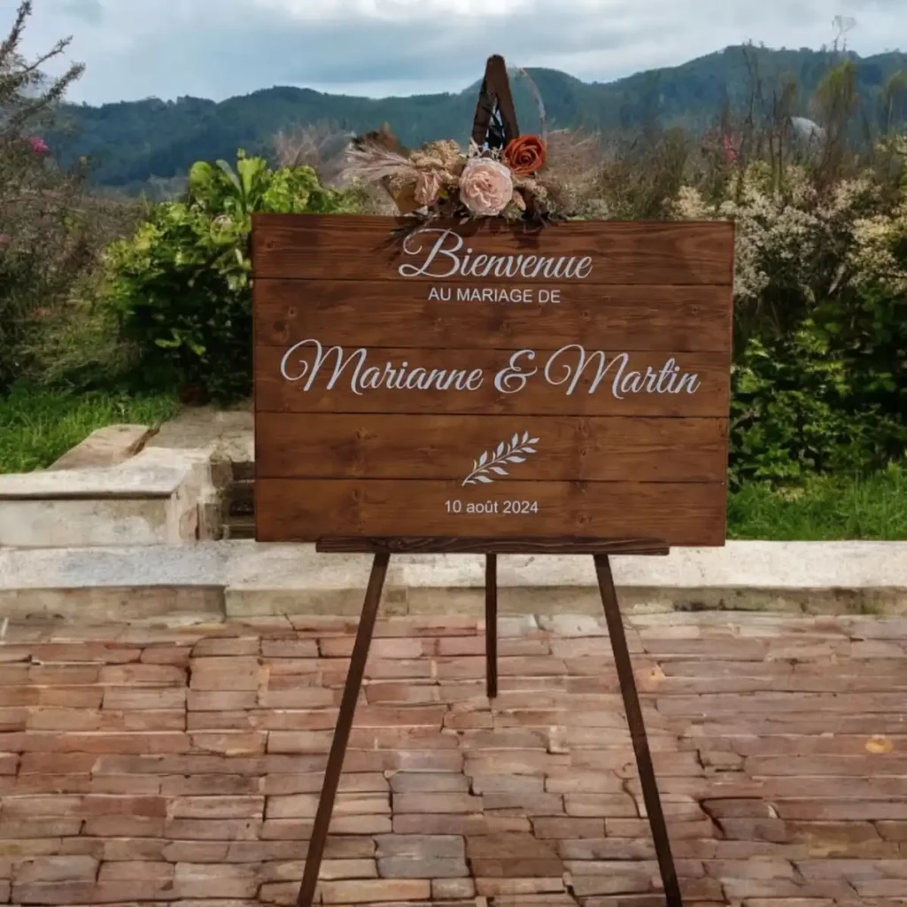 Location panneau de bienvenue sur mesure - Panneau de bienvenue personnalisé en bois pour un mariage, affichant les noms des mariés, Marianne et Martin, et la date du 10 août 2024. Le panneau est décoré de fleurs artificielles et repose sur un trépied en bois, placé dans un cadre extérieur pittoresque avec des collines verdoyantes en arrière-plan. Ce panneau rustique et élégant est parfait pour accueillir les invités à un mariage champêtre et personnalisé.