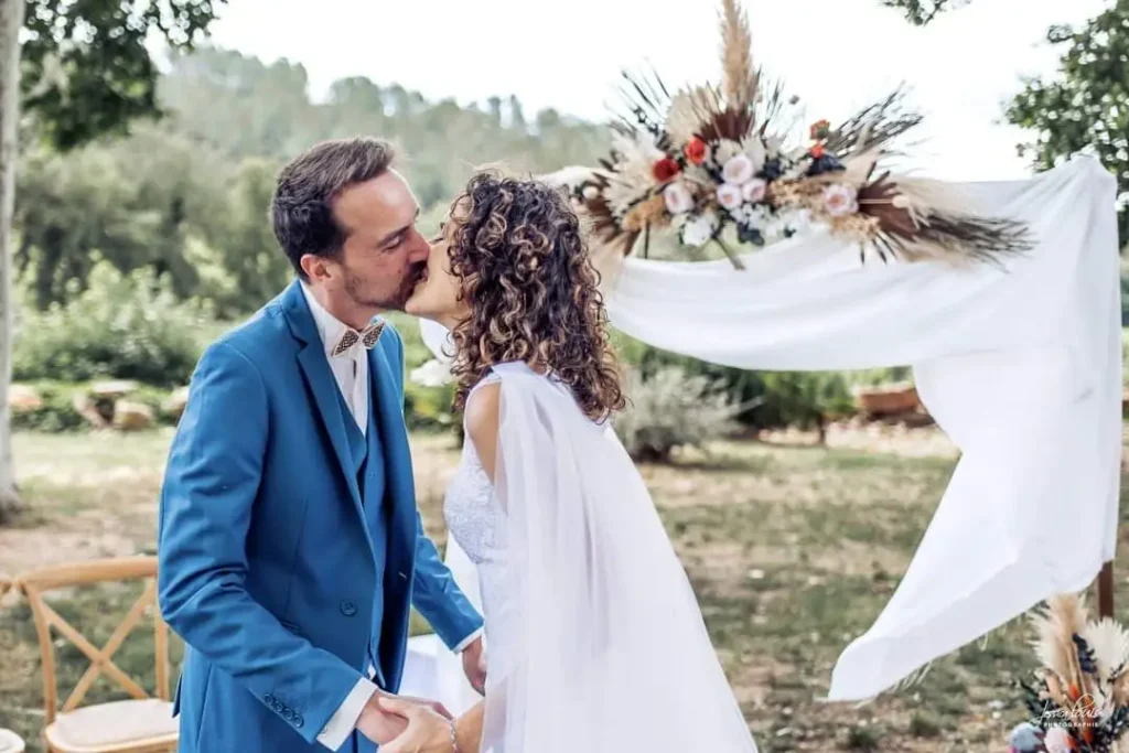 location arches fleurs et voilages en location - Couple de mariés s'embrassant sous une arche de mariage décorée de fleurs et de voilages blancs. Le marié porte un costume bleu vif avec un nœud papillon beige, et la mariée une robe blanche en dentelle avec une cape fluide. L'arche est ornée de pampas, de roses et de feuillage, créant un cadre romantique en plein air pour la cérémonie.