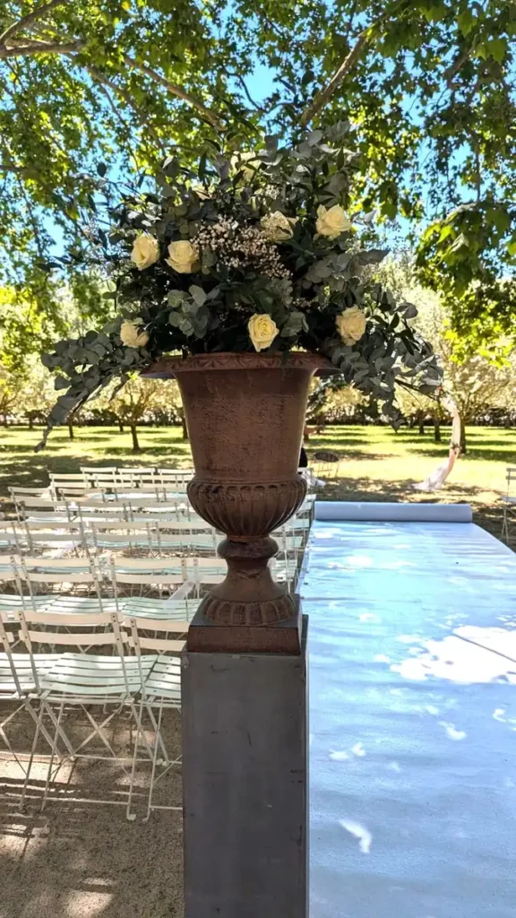 Grande composition florale de roses blanches et de feuillage vert dans un vase Médicis en fer forgé, située à l'entrée d'une allée de mariage en plein air. Les chaises blanches sont disposées pour les invités sous un couvert d'arbres, créant une ambiance ombragée et élégante pour la cérémonie. L'allée centrale est recouverte d'un tapis blanc, menant à une arche décorative.