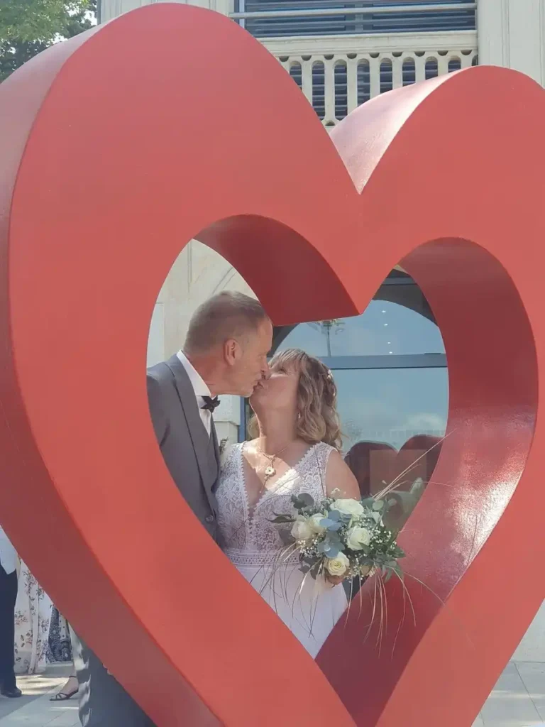 Création bouquet de mariage dans le Var - Un couple de mariés s'embrassant à travers une sculpture de cœur rouge. La mariée porte une robe en dentelle blanche et tient un bouquet de fleurs blanches et vertes, tandis que le marié porte un costume gris avec un nœud papillon noir. La scène est capturée devant un bâtiment avec une architecture élégante