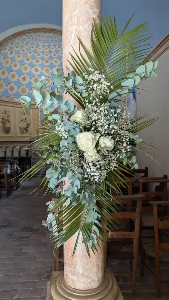 Arrangement floral pour mariage attaché à une colonne en pierre, composé de roses blanches, de gypsophiles et de feuillage vert, incluant des feuilles de palmier et de l'eucalyptus. L'intérieur de l'église est visible en arrière-plan, avec des bancs en bois et des fresques murales, créant une atmosphère élégante et sacrée pour la cérémonie de mariage.