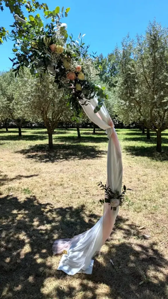 création bouquet de fleurs fraîches pour arche de mariés - Arche de mariage en plein air décorée de fleurs fraîches, incluant des roses blanches et roses, du gypsophile et du feuillage vert. L'arche est drapée de tissu blanc et rose pâle, créant une ambiance romantique et élégante au milieu d'un verger d'oliviers. Parfait pour une cérémonie de mariage champêtre et naturelle.