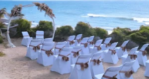 Cette photo capture une cérémonie de mariage en bord de mer, avec des chaises blanches soigneusement alignées sur le sable et ornées de rubans marron. Une allée centrale conduit à une arche décorée de fleurs séchées et de feuillage, située juste devant l'océan. À l'arrière-plan, on peut apercevoir des rochers et quelques bateaux au loin, avec des collines en arrière-plan sous un ciel bleu clair. La scène dégage une atmosphère paisible et romantique, parfaite pour une célébration en extérieur au bord de l'eau.