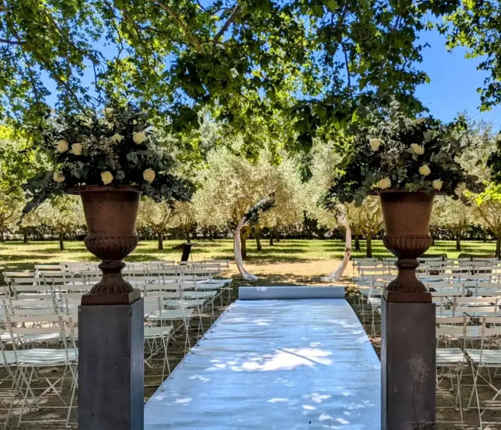 composition florale pour arche et vases medicis - Cérémonie de mariage en plein air avec une allée centrale bordée de grands vases Médicis contenant des compositions florales de roses blanches et de feuillage vert. Des chaises blanches sont disposées pour les invités sous un couvert d'arbres, créant une ambiance ombragée et sereine. Une arche décorative se trouve au bout de l'allée, ajoutant une touche élégante à la scène.