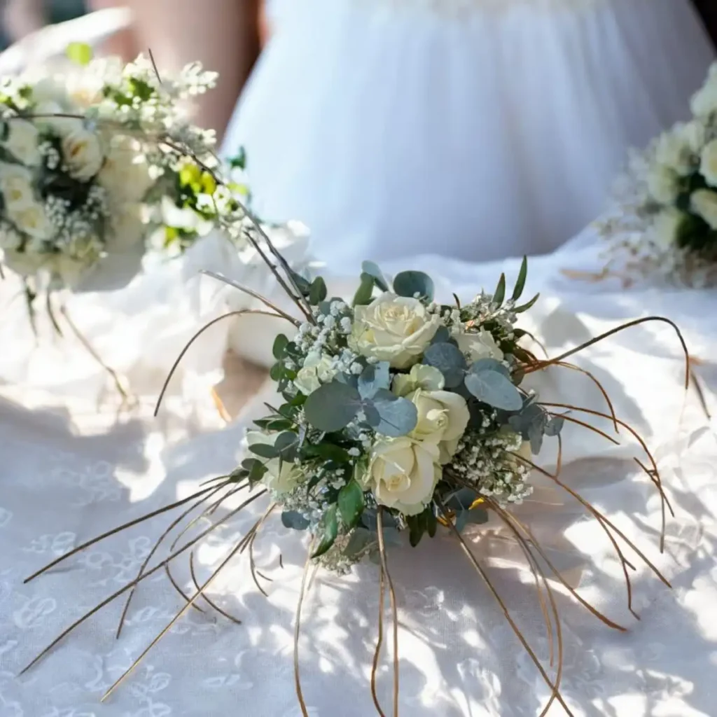 Création bouquet de marié dans le var - Bouquet de mariage en fleurs fraîches composé de roses blanches, de gypsophiles et de feuillage vert, posé sur une nappe blanche en dentelle. Le bouquet est élégamment arrangé avec des branches décoratives, créant un look romantique et naturel pour la mariée.
