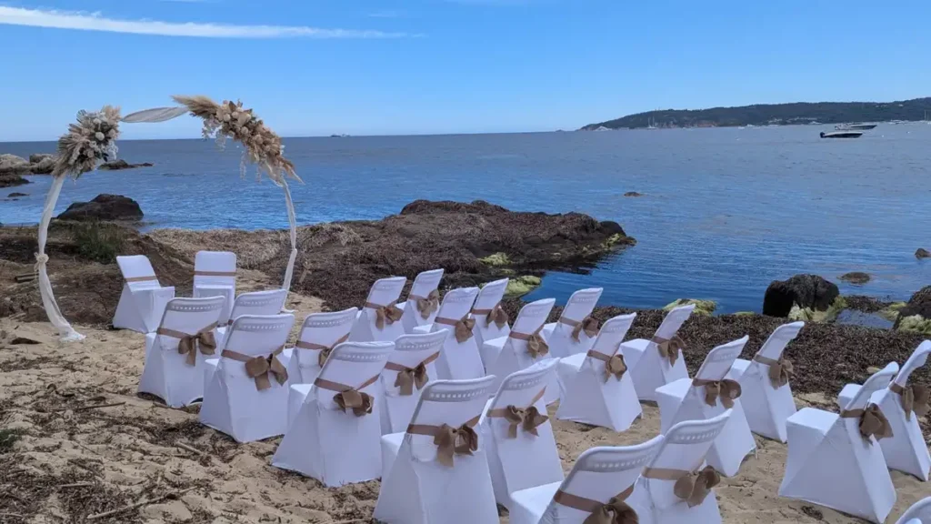 Décoration florale arche en location - Cette photo montre une cérémonie laïque de mariage organisée sur une plage pittoresque. Des chaises blanches élégamment décorées avec des nœuds marron clair sont disposées en rangées, tournées vers une arche florale décorée de fleurs séchées et de rubans blancs, face à l'océan. En arrière-plan, on peut voir les vagues de la mer et une étendue de verdure ajoutant une touche naturelle et sereine à l'ensemble. Le ciel est dégagé et bleu, offrant une journée parfaite pour une célébration en extérieur.