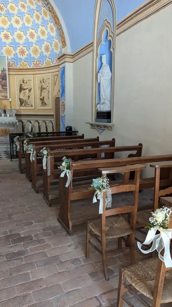 gerbes de feurs sur banc pour mariage - Bancs en bois décorés de bouquets de fleurs fraîches attachés avec des rubans blancs, disposés dans une église ornée de fresques et de statues pour une cérémonie de mariage. L'intérieur de l'église présente un sol en briques et des murs richement décorés, créant une atmosphère sacrée et élégante pour les noces.