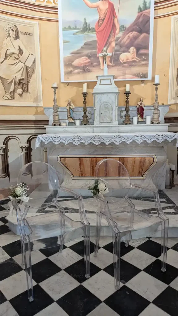 Chaises transparentes décorées de petites gerbes de fleurs blanches et de feuillage, disposées devant l'autel d'une église pour une cérémonie de mariage. L'autel est orné de statues religieuses, de chandeliers et de napperons en dentelle, avec un sol en damier noir et blanc créant une ambiance élégante et sacrée.