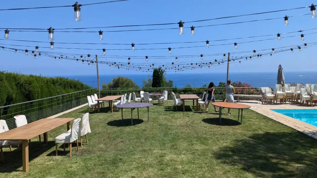 installation de guirlandes guinguettes dans le var - Installation guinguette pour un événement en plein air dans le Var, avec des tables et des chaises disposées sur une pelouse verdoyante. Des guirlandes lumineuses suspendues au-dessus offrent une ambiance festive et romantique. La vue panoramique sur la mer Méditerranée ajoute une touche spectaculaire à l'ensemble, créant un cadre parfait pour une réception en extérieur.
