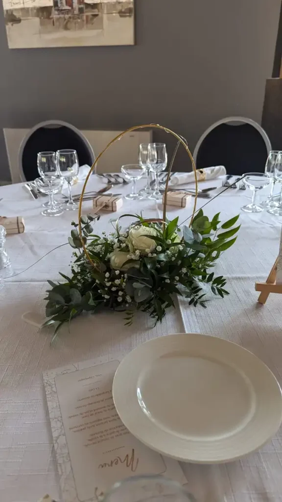 Location centre de table pour un mariage au kouros cuers - Centre de table de mariage élégant avec des fleurs blanches et des feuilles vertes, disposé sur une table dressée avec de la vaisselle et des verres à vin. La décoration florale est placée dans un cercle doré, ajoutant une touche raffinée à la table. Menu de mariage et assiette blanche visibles au premier plan, offrant une ambiance sophistiquée pour un événement spécial.