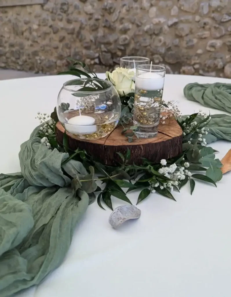 location de centre de table, création sur mesure en rondin et fleurs fraîches - Centre de table de mariage rustique composé d'un rondin de bois, de bougies flottantes dans des vases en verre, et de fleurs fraîches blanches entourées de feuillage vert. Des tissus verts drapés ajoutent une touche d'élégance naturelle à la décoration, idéale pour une réception de mariage champêtre.