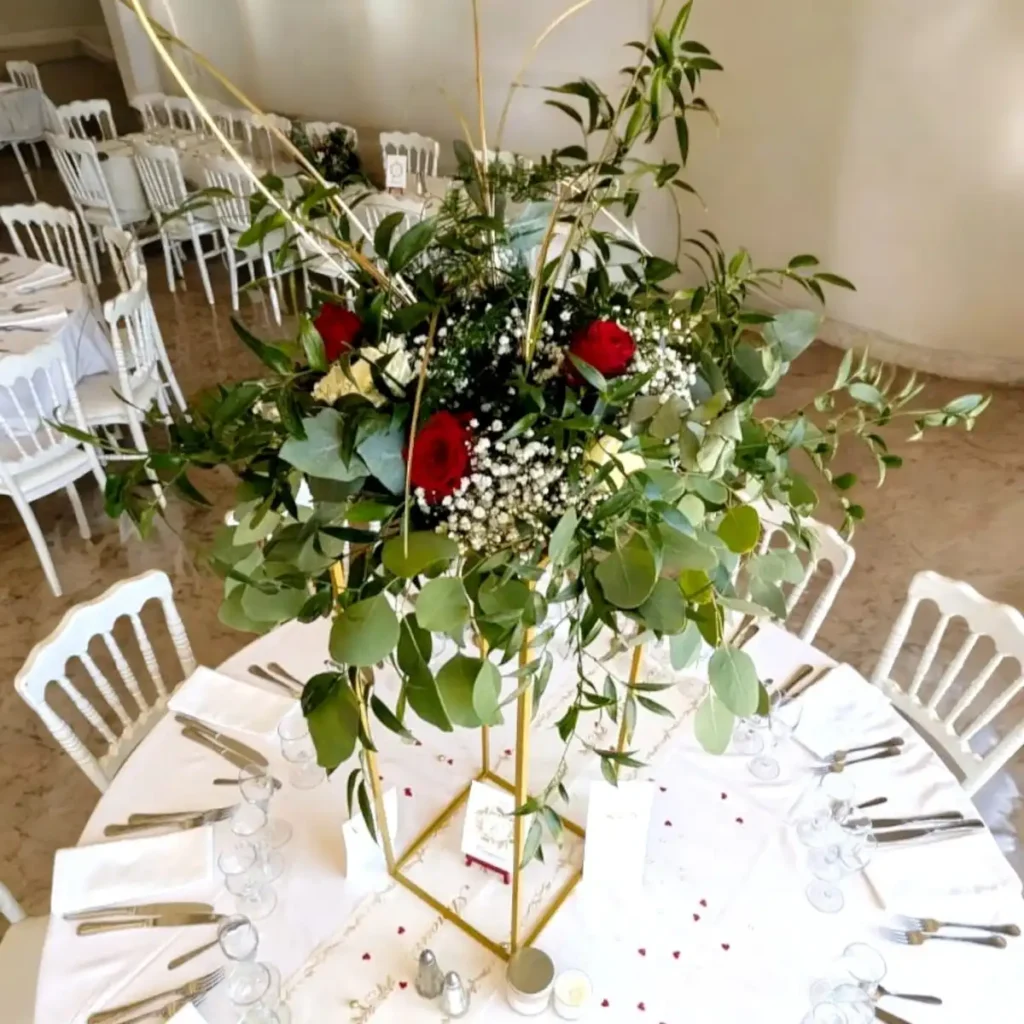 Centre de table floral élégant pour un mariage dans le Var, composé de roses rouges et blanches, de gypsophile et de feuillage vert, disposé sur un support doré. La décoration de table est complétée par des chaises blanches et une nappe blanche, créant une ambiance raffinée et romantique pour la réception. Idéal pour un mariage à thème en rouge et blanc avec des fleurs fraîches.