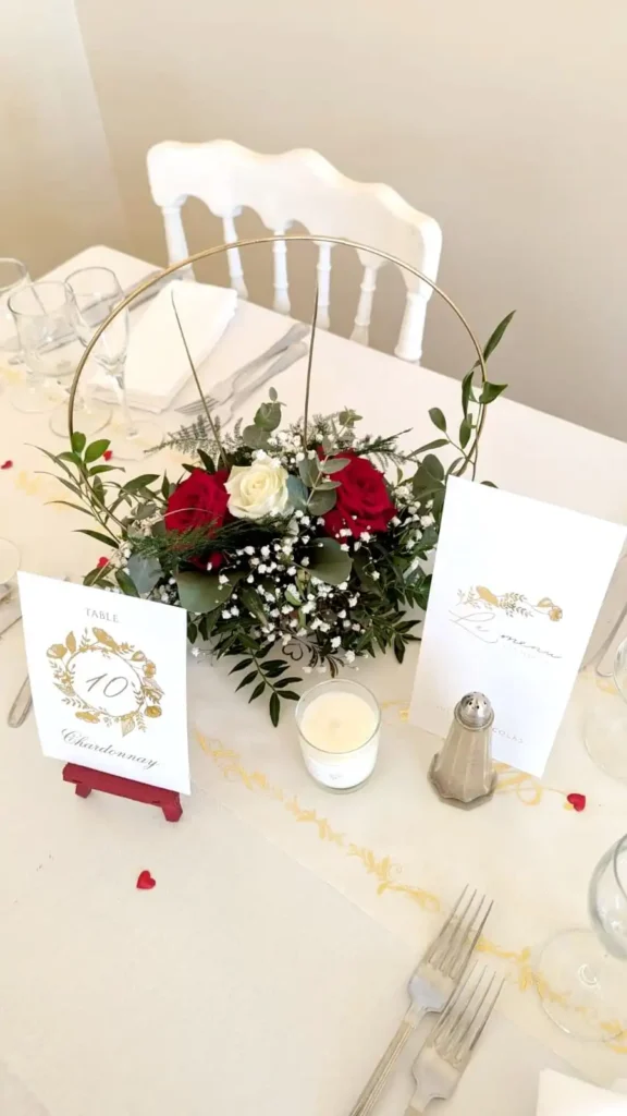 Centre de table floral pour mariage avec des roses rouges et blanches, accompagné de gypsophile et de feuillage, présenté dans un cercle doré sur une table élégante. La décoration comprend également une carte de menu et une carte de numéro de table, marquée 'Table 10 Chardonnay', le tout sur une nappe blanche avec une ambiance raffinée et romantique. Idéal pour un mariage dans le Var avec un thème classique et floral.