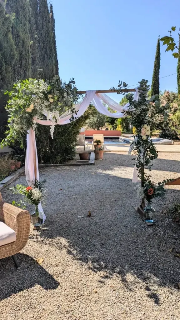 Installation de l'arche de mariage carrée en bois décorée de fleurs blanches et de verdure au Clos Céline à Flayosc, disponible à la location chez Iliade Location. Cadre romantique avec chaises et piscine en arrière-plan, parfait pour une cérémonie en plein air.