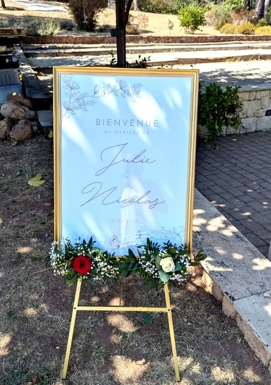 Panneau de bienvenue au mariage de Julie et Nicolas, daté du 10 août 2024, placé à l'extérieur sur un chevalet doré. Le panneau est décoré avec une guirlande florale composée de roses rouges et blanches, de gypsophile et de feuillage, dans un cadre champêtre dans le Var. Idéal pour un accueil chaleureux des invités à la cérémonie de mariage.