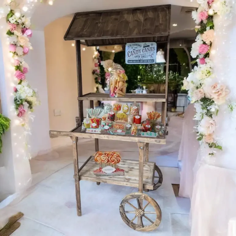 Un charmant chariot en bois de style vintage, décoré pour un "Candy Bar", est placé dans un espace intérieur élégant, encadré par des colonnes de fleurs blanches et roses, agrémentées de guirlandes lumineuses. Le chariot est rempli de divers bonbons colorés, présentés dans des bocaux et sur des plateaux. Des sucettes géantes décorent l'étagère inférieure, tandis qu'une enseigne rétro indiquant "Candy Canes" est accrochée sous le toit en bois. L'arrière-plan est un décor festif et romantique, avec des détails floraux lumineux.