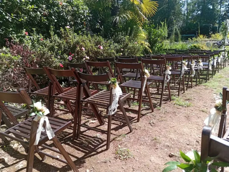 Des rangées de chaises en bois sont alignées en plein air pour une cérémonie laïque, chaque chaise étant décorée d'un petit bouquet de fleurs blanches et de feuillage vert, attaché avec un ruban blanc transparent. L'installation se trouve dans un jardin verdoyant, avec des buissons fleuris en arrière-plan et une ambiance naturelle et paisible, créant un cadre romantique et rustique pour la cérémonie.