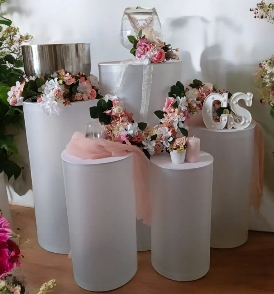 Colonne cylindrique de mariage : Arrangement de décoration de mariage avec des colonnes blanches et transparentes. Les colonnes sont décorées de fleurs artificielles dans des tons de rose, blanc et vert. Des bougies roses et une écharpe en tulle rose ajoutent une touche romantique. Un grand vase en verre et des lettres décoratives "CS" sont également présents sur les colonnes.