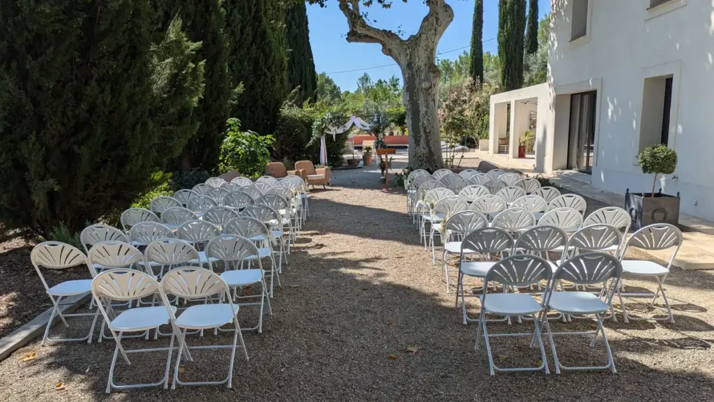 Disposition de chaises blanches pliantes en plein air pour une cérémonie laïque, formant une allée centrale sous un grand arbre, avec un autel en arrière-plan.