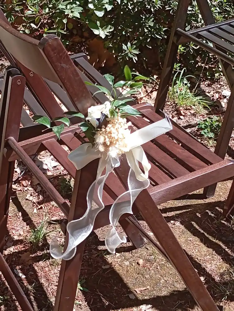 Un gros plan d'une chaise en bois pliable utilisée pour une cérémonie laïque en plein air, décorée d'un petit bouquet de fleurs blanches et de feuillage vert, attaché à l'accoudoir avec un ruban blanc transparent qui se drape élégamment. En arrière-plan, on aperçoit des plantes et des feuilles au sol, créant une ambiance naturelle et rustique.