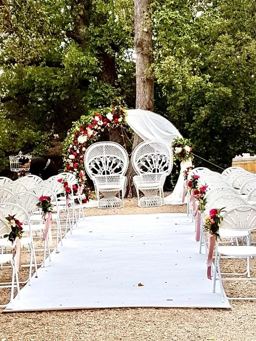 Cette image montre une disposition élégante pour une cérémonie de mariage en plein air. Un tapis blanc est posé au centre, créant une allée qui mène à deux grands fauteuils décoratifs en osier blanc, placés sous un bel arc fleuri. L'arc est orné de fleurs rouges et blanches, avec des touches de verdure, ajoutant une touche romantique à la scène. Les chaises blanches en fer forgé, alignées de part et d'autre de l'allée, sont décorées de petits bouquets de fleurs rouges et blanches, maintenus par des rubans roses. L'ensemble est situé dans un cadre naturel avec des arbres imposants en arrière-plan, offrant une atmosphère à la fois intime et majestueuse pour une cérémonie de mariage.