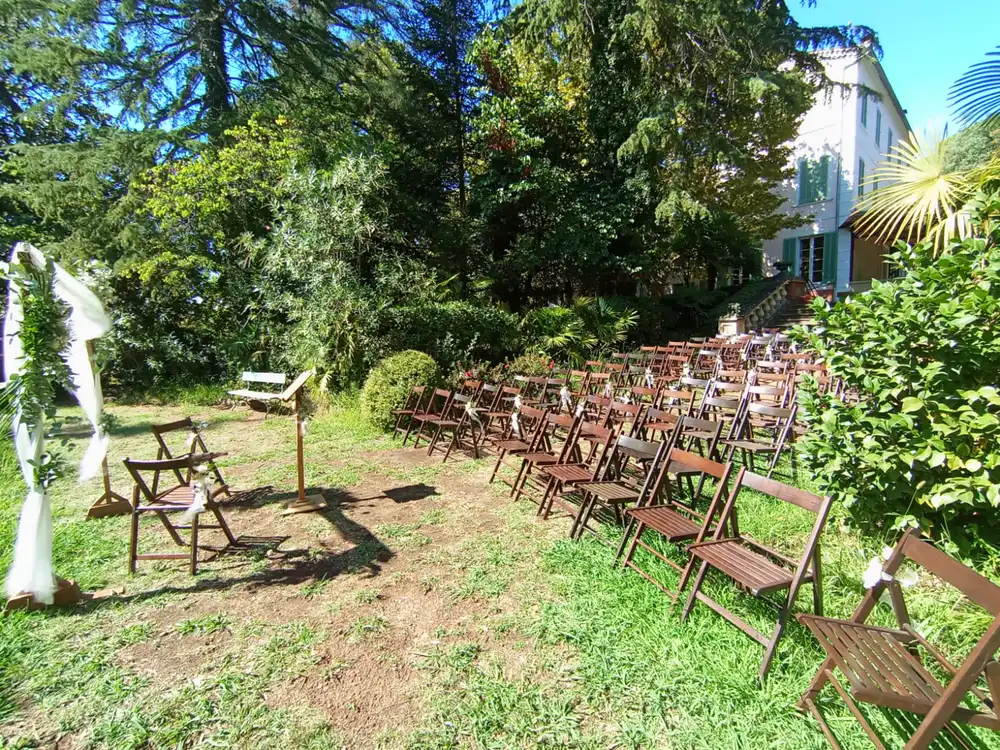 Une installation en plein air pour une cérémonie laïque avec des rangées de chaises en bois disposées sur une pelouse verdoyante. À gauche de l'image, on aperçoit une arche décorée de tissu blanc et de verdure, ainsi qu'un pupitre en bois. Les chaises font face à l'arche, prêtes à accueillir les invités. En arrière-plan, un grand bâtiment blanc avec des volets verts est entouré d'arbres luxuriants et de buissons, créant un cadre naturel et paisible pour l'événement.