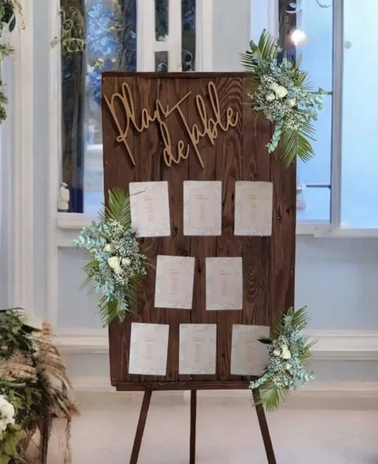 Panneau en bois de plan de table de mariage, décoré de feuillages verts et de petites fleurs blanches. Le panneau est orné du texte 'Plan de Table' en écriture cursive en bois doré, et présente des cartes indiquant les noms des invités et leur placement à table, affichées dans un style épuré et élégant.