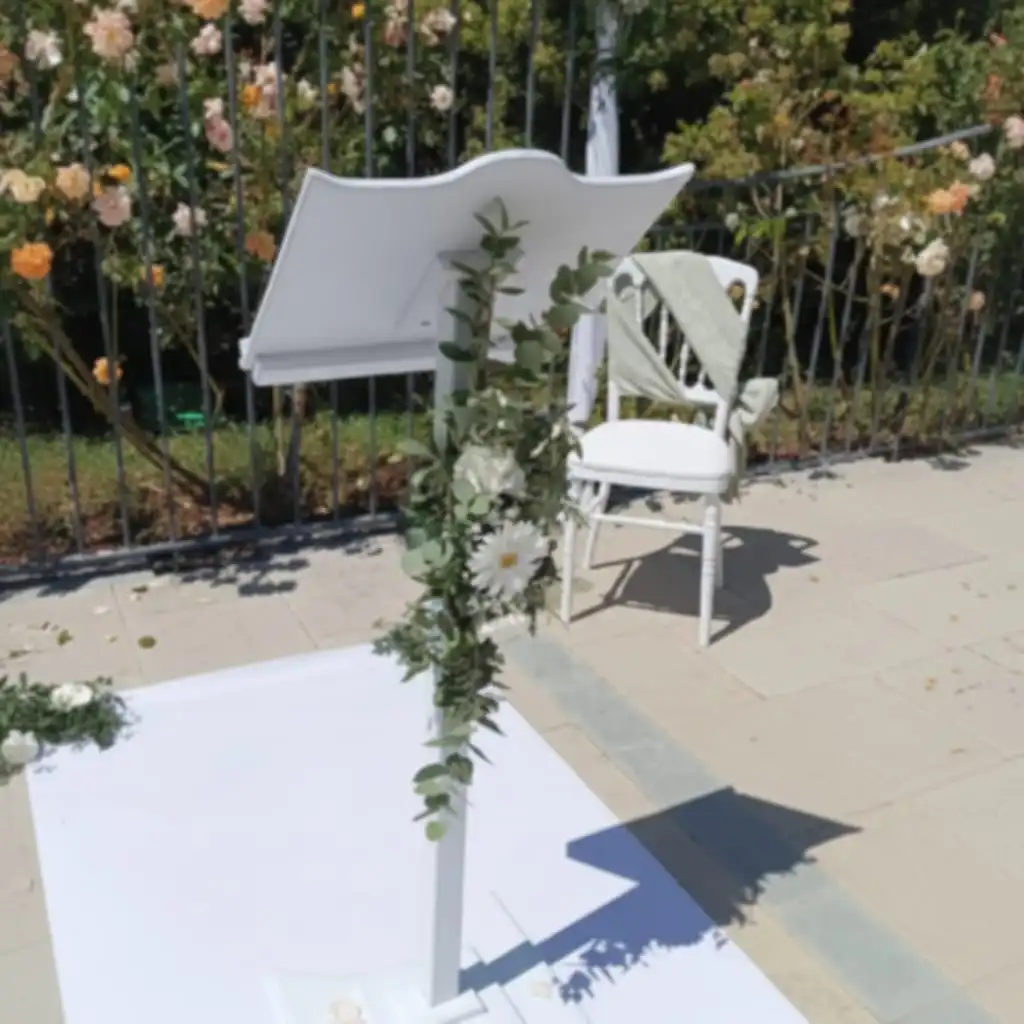 Pupitre de cérémonie de mariage en bois blanc décoré de feuillages et de fleurs blanches, installé en extérieur devant un jardin fleuri.