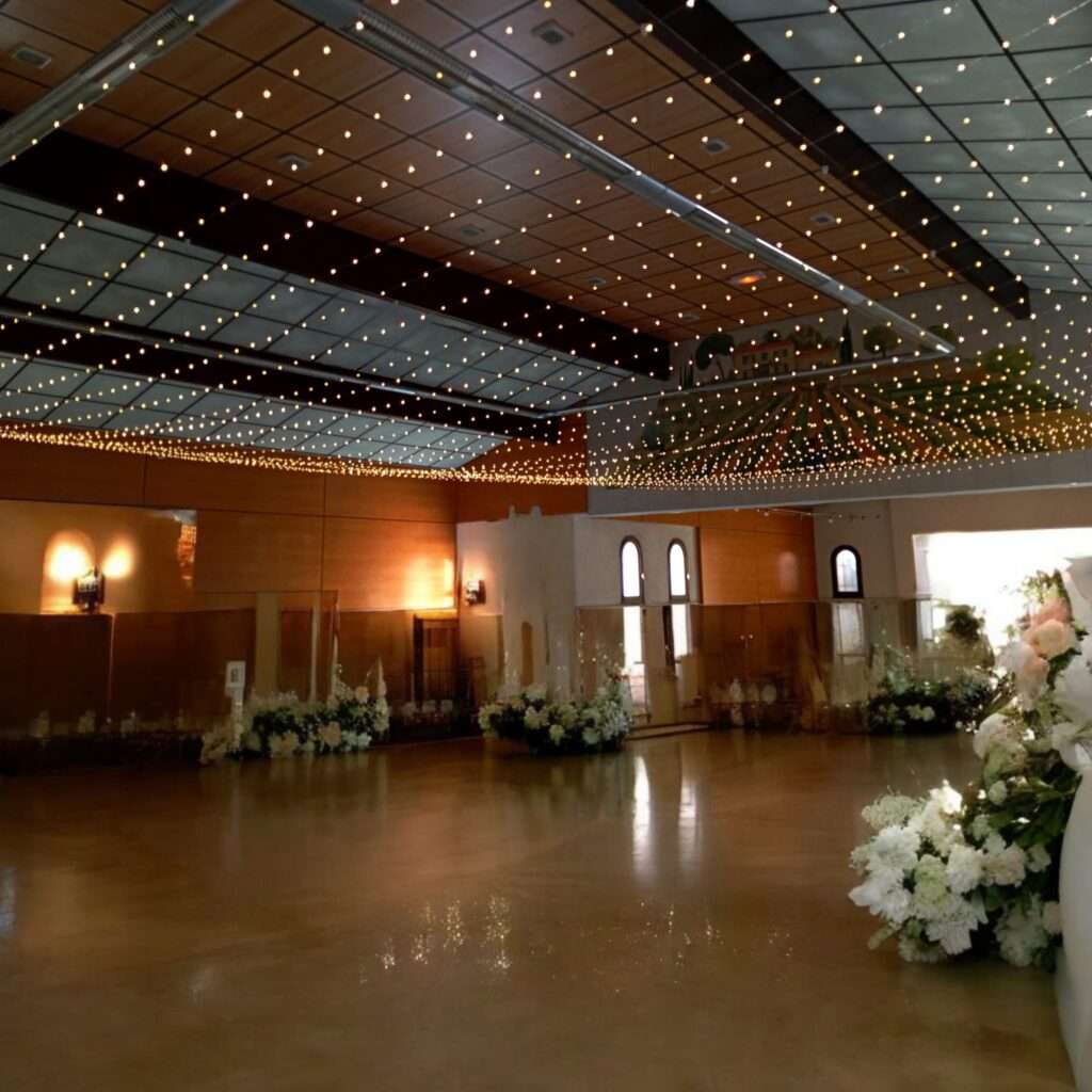 Salle de réception de mariage avec un plafond illuminé de guirlandes lumineuses créant un effet de ciel étoilé, décorée de fleurs blanches le long des murs et d'éléments en bois, offrant une ambiance chaleureuse et élégante.
