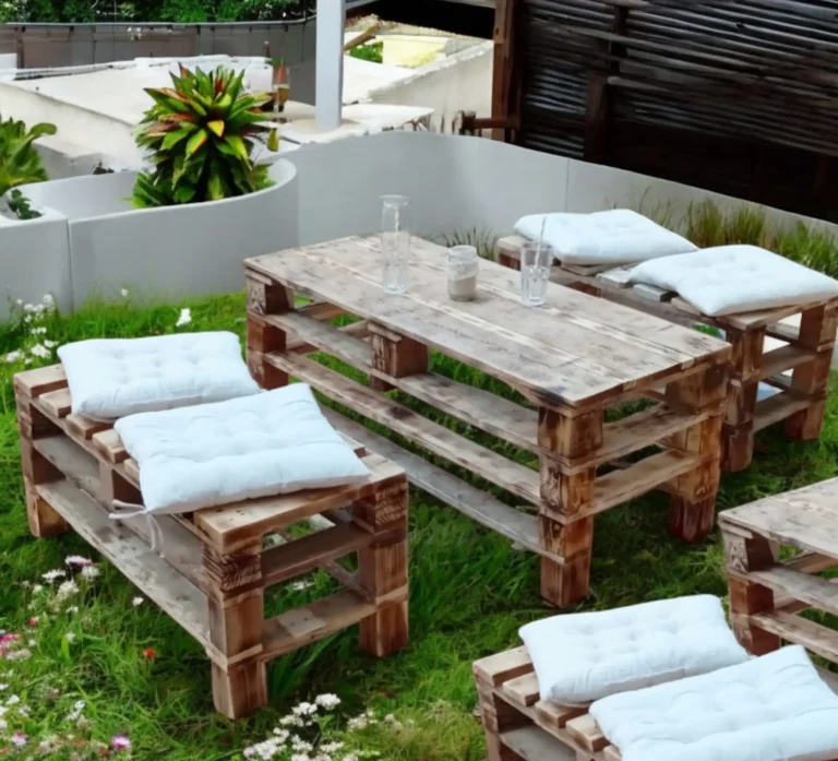 Location de salon de jardin : Salon de jardin en bois de palette avec coussins blancs, installé dans un jardin fleuri, parfait pour une ambiance champêtre et bohème lors d'un mariage ou d'un événement extérieur.