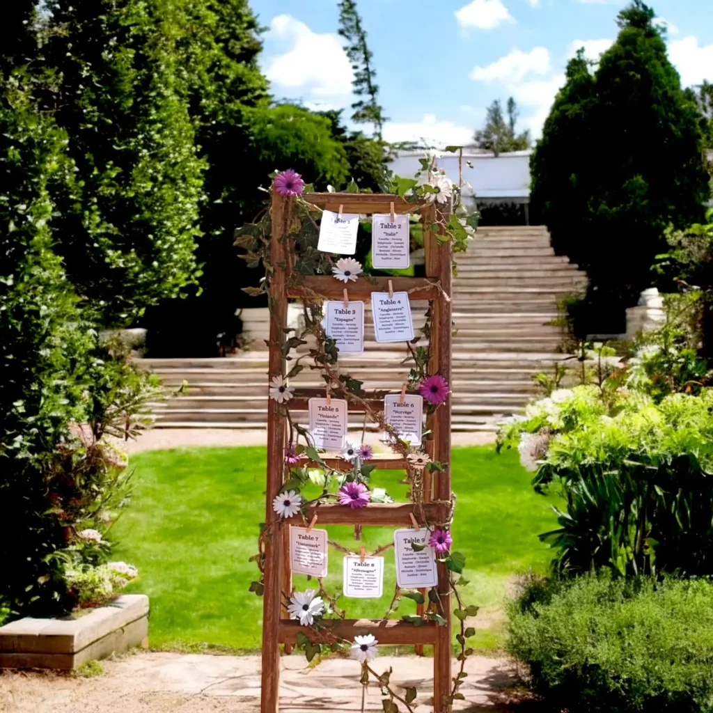 Échelle en bois transformée en plan de table de mariage, ornée de fleurs champêtres et de feuillage, avec des cartons nominatifs suspendus à l’aide de pinces à linge, placée dans un jardin élégant face à un grand escalier en pierre.