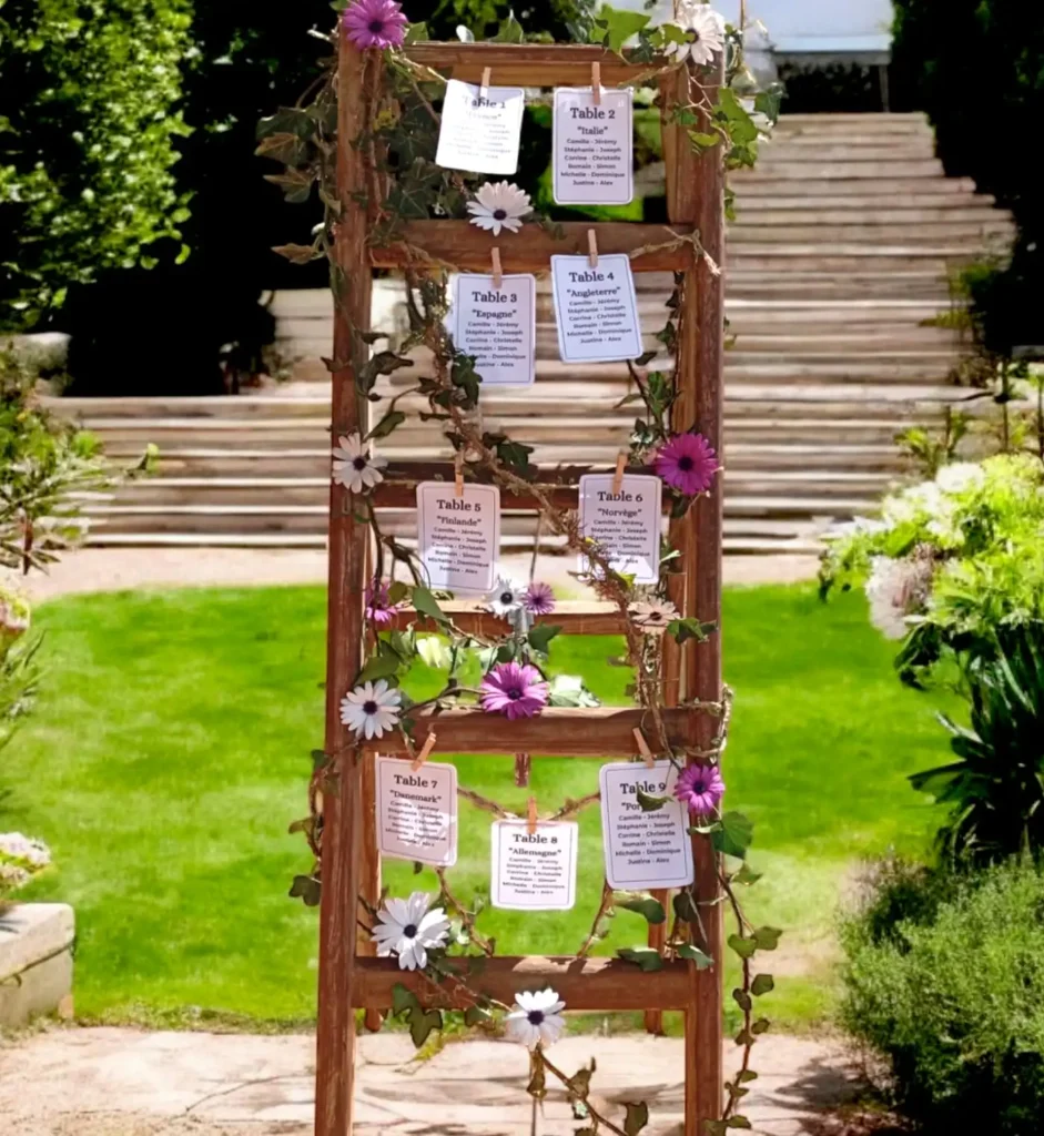 Panneau de plan de table en bois décoré de fleurs et de feuillage, affichant les noms des invités par table pour un mariage en extérieur, placé dans un jardin verdoyant avec escaliers en arrière-plan