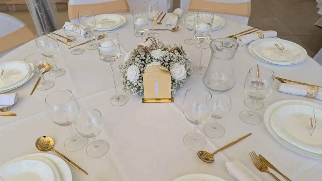 Table de mariage élégante décorée en blanc et or, centre de table fleuri avec gypsophile et roses blanches, numéro de table doré, vaisselle raffinée et couverts dorés.