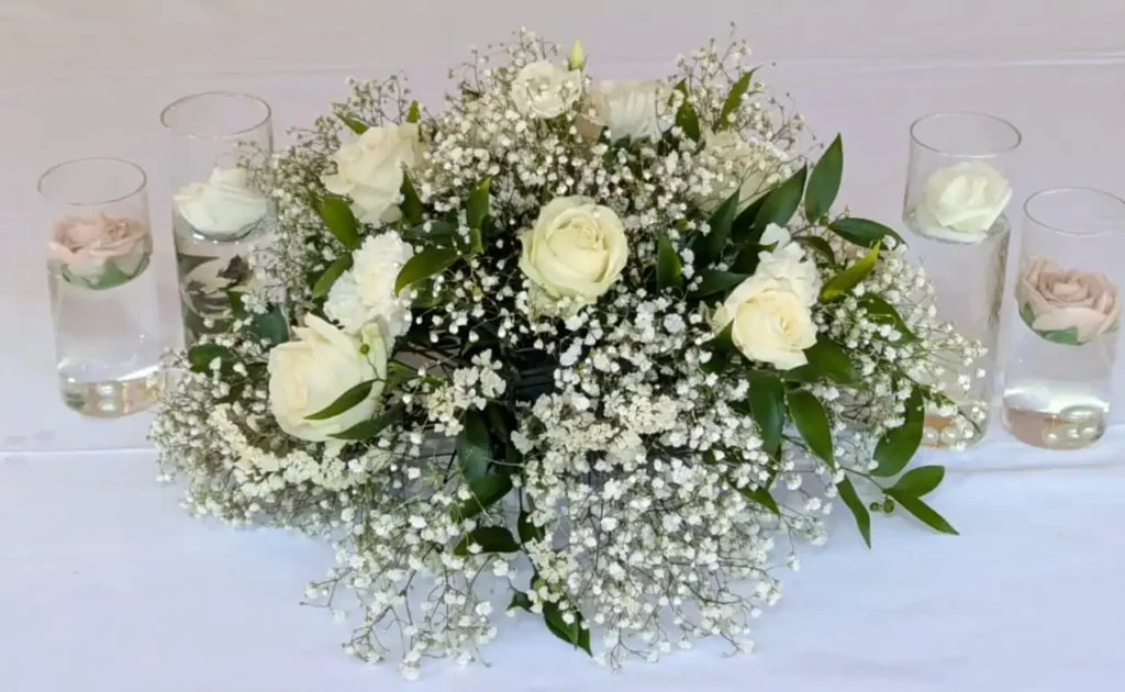 Centre de table de mariage composé de roses blanches, de gypsophile et de feuillage vert, entouré de verres cylindriques contenant des roses flottantes dans l’eau.