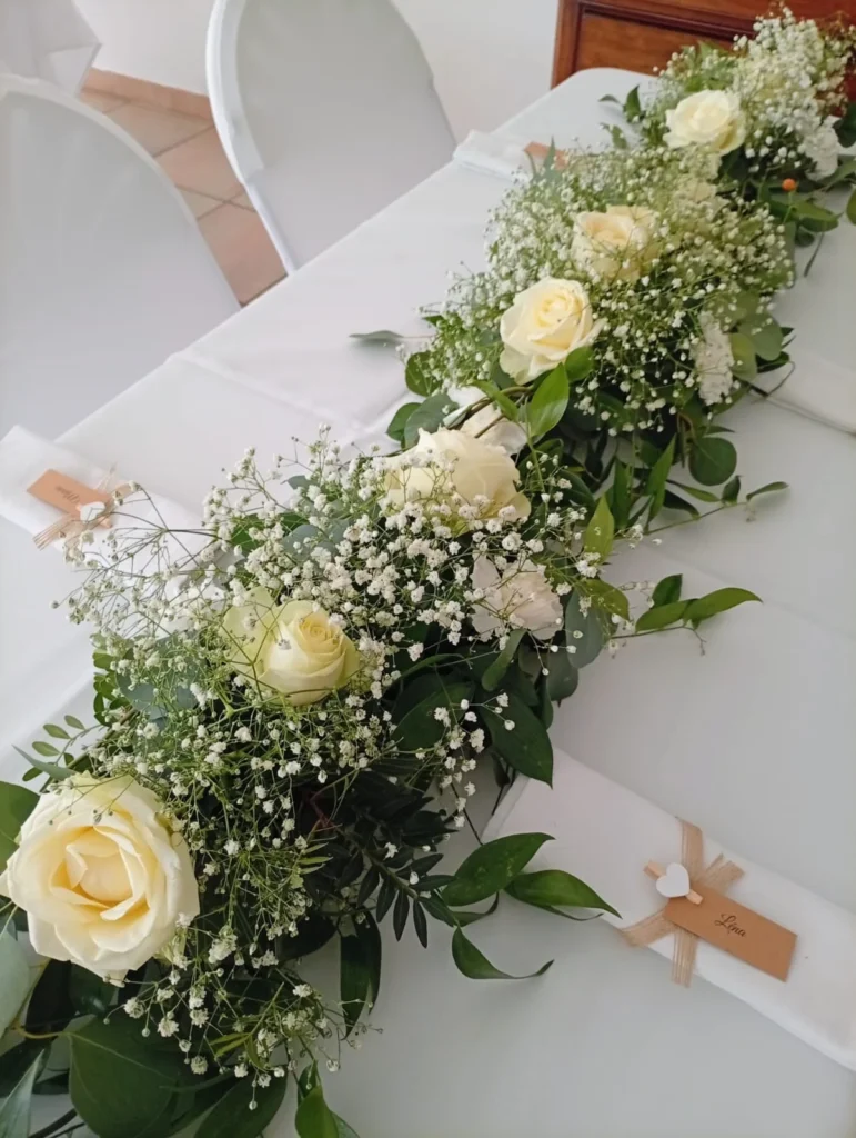 Centre de table de mariage avec une guirlande florale composée de roses blanches, gypsophile et ruscus sur une nappe blanche.