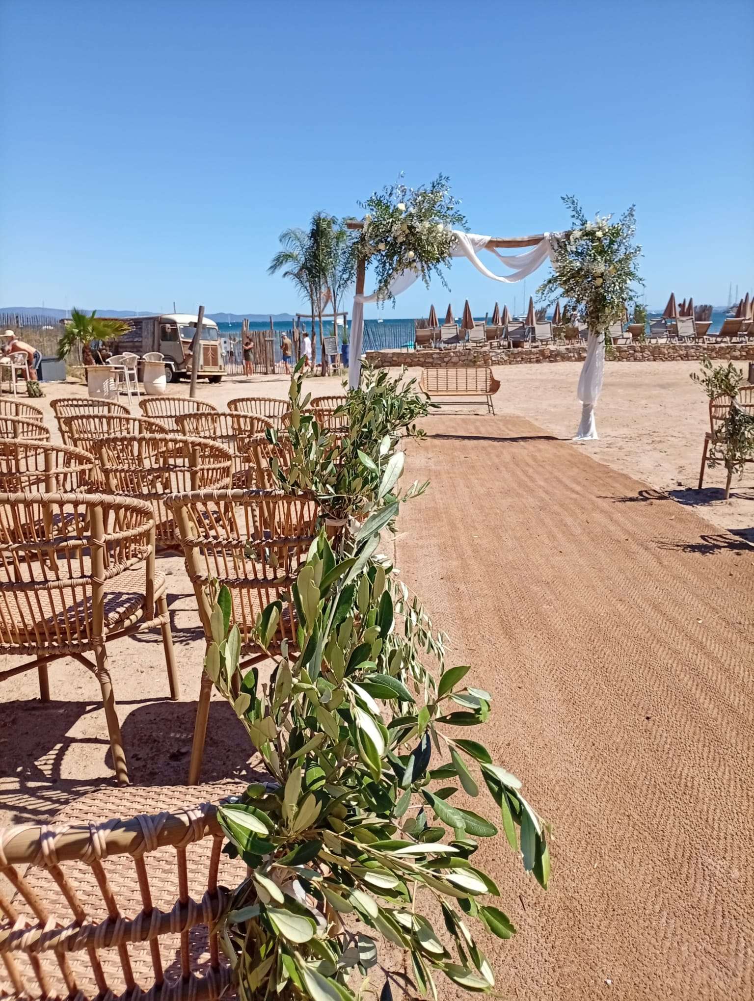Allée de cérémonie sur sable pour mariage en bord de mer, chaises en rotin tressé, arche fleurie et feuillage d'olivier.