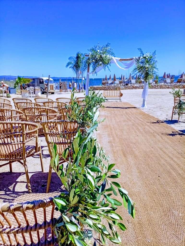 Arche de mariage décorée de feuillage d’olivier et de voilage blanc, sur fond de plage et de transats en bord de mer au Noé plage à Hyères 83400