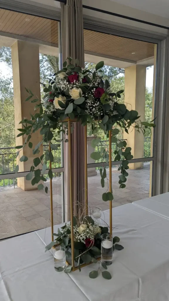 Centre de table de mariage sur colonne dorée, décoré de roses blanches et rouges, feuillage d’eucalyptus et gypsophile, pour un décor de réception raffiné et lumineux.