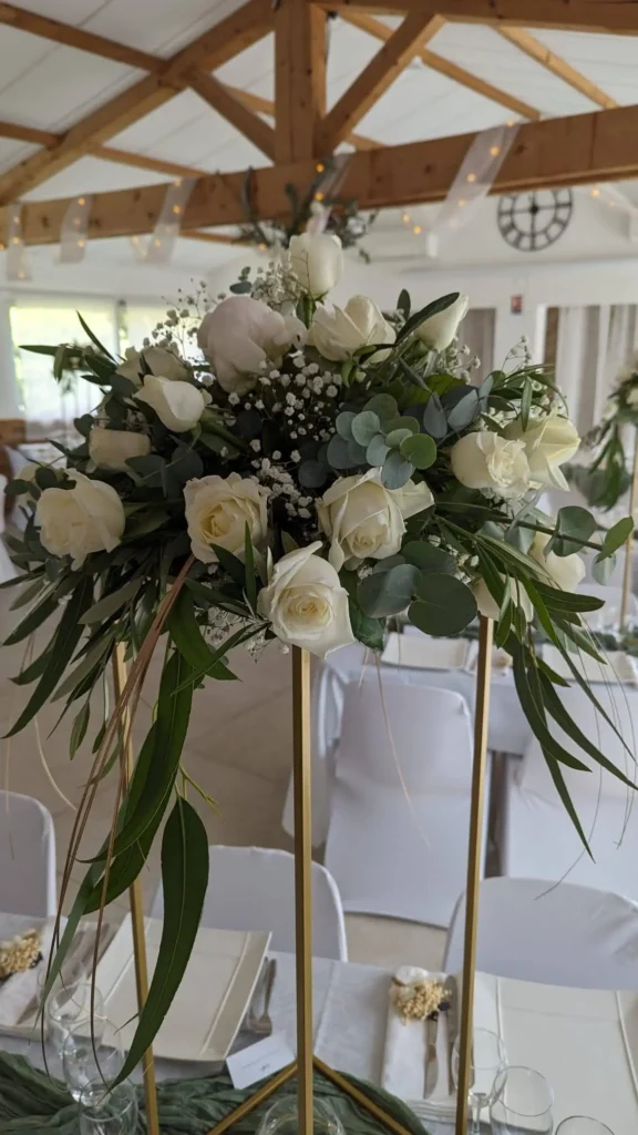 Grand centre de table floral sur structure dorée avec bouquet de roses blanches, eucalyptus et gypsophile. Décoration de mariage naturelle et luxueuse, feuillage retombant, ambiance romantique et verdoyante sous charpente en bois, tables nappées de blanc et décoration raffinée.