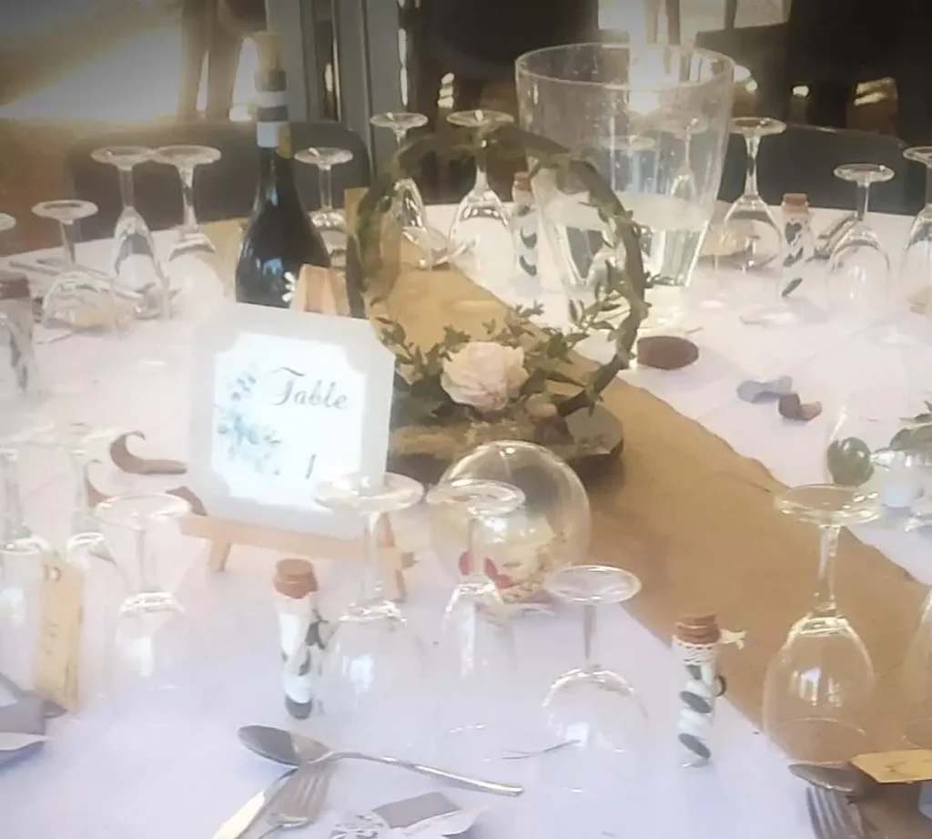 Décoration de table de mariage rustique et champêtre avec chemin de table en toile de jute, vaisselle transparente, verres retournés et rondins de bois. Centre de table composé d’un cercle végétal orné de feuillage, lierre et fleur rose poudré, ambiance naturelle douce et authentique pour réception champêtre élégante.