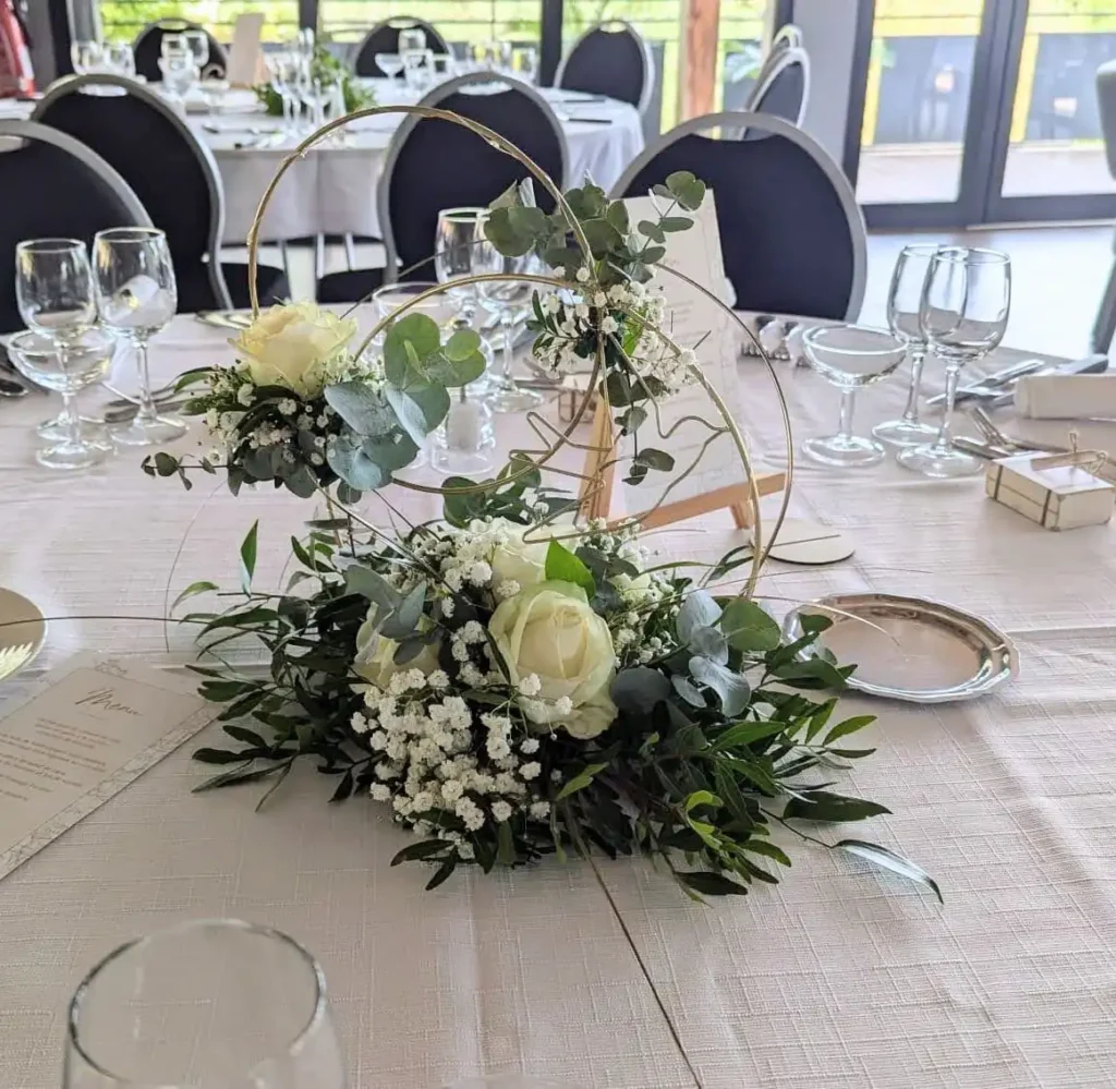 Centre de table rond pour mariage, composition florale moderne avec cercles dorés, roses blanches, eucalyptus et gypsophile. Disposition élégante sur nappe ivoire, ambiance romantique et contemporaine, parfaite pour un mariage nature chic ou bohème.
