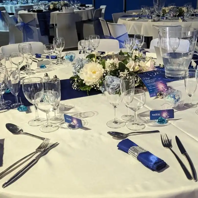Table de mariage bleu marine et blanche raffinée, nappe immaculée, serviettes satinées bleu roi, porte-noms personnalisés, composition florale centrale avec pivoines et roses blanches, gypsophile et feuillage vert tendre. Ambiance chic et romantique pour réception élégante en salle.