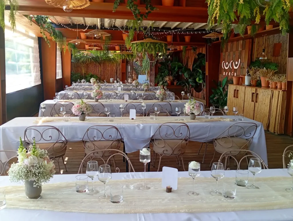 Salle de réception de mariage bohème et naturelle décorée de tables rectangulaires avec centres de table fleuris, vases en grès, gypsophile, roses pastel et feuillage. Ambiance tropicale et chaleureuse avec suspension en rotin, bois, bambou et plantes suspendues, esprit guinguette chic et nature.