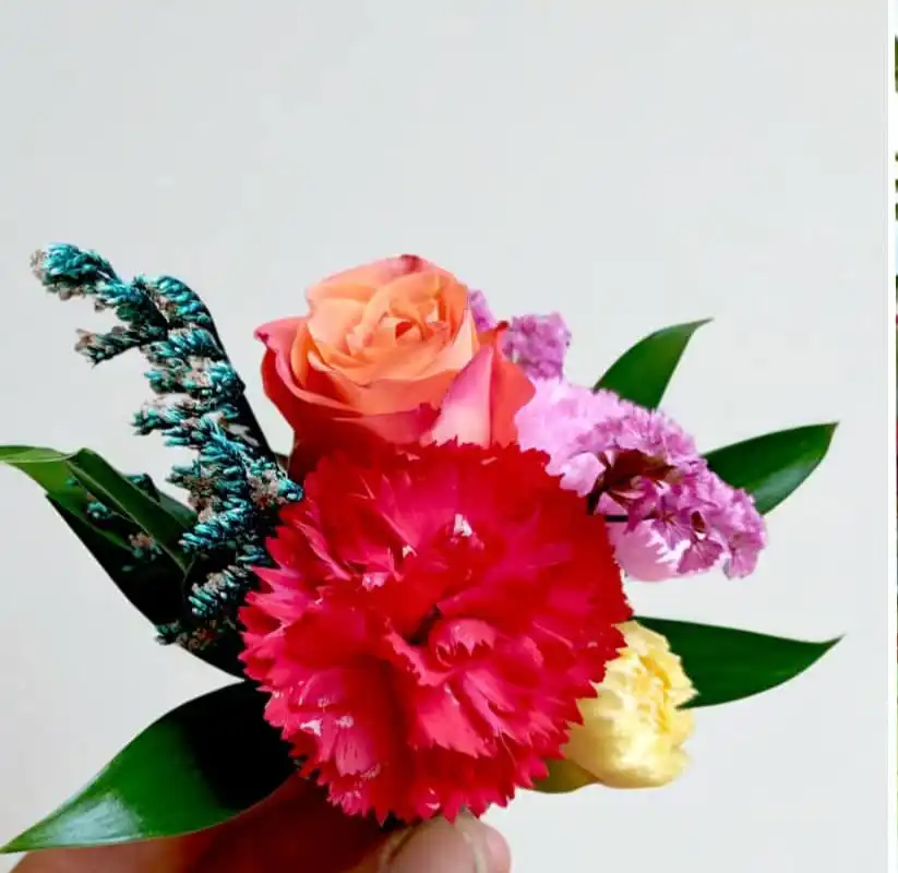 Boutonnière de mariage colorée avec rose orange, œillet rouge, fleurs mauves et feuillage vert, tenue à la main sur fond clair.