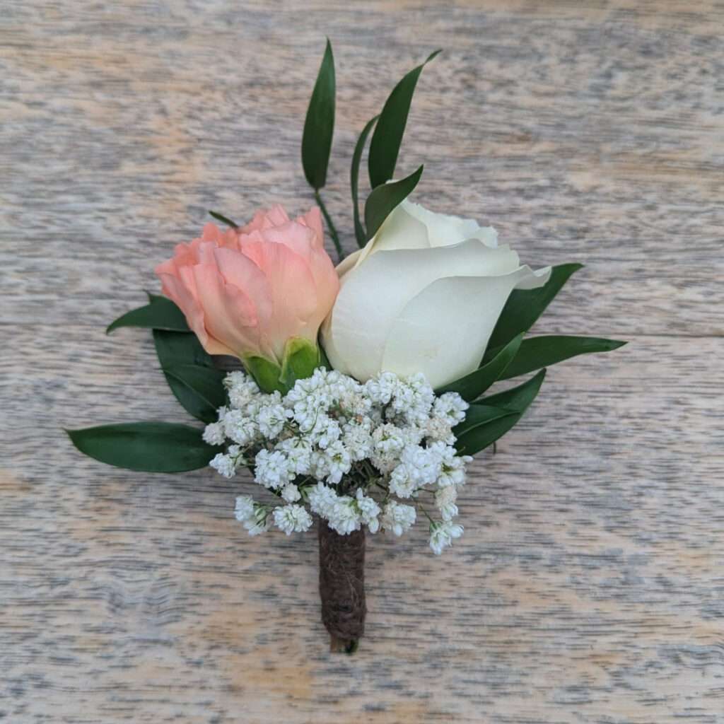 Boutonnière de mariage composée d’une rose blanche, d’une rose pêche, de gypsophile blanc et de feuillage vert, montée sur tige et posée sur une table en bois.