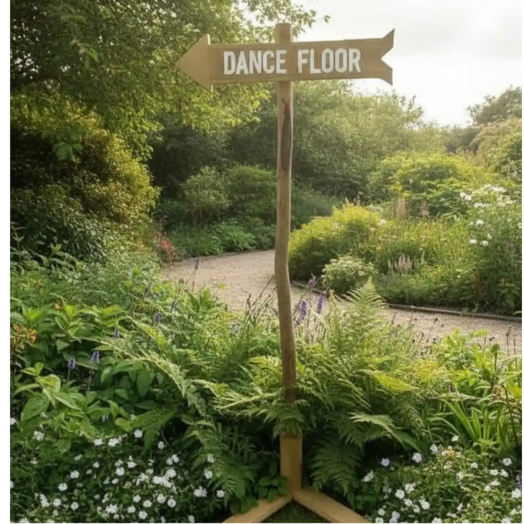 Flèche directionnelle en bois naturel indiquant “Dance Floor”, montée sur un pied en rondin et installée dans un jardin de mariage au décor champêtre.