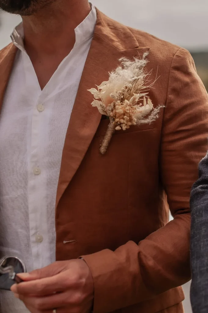 Boutonnière de marié en fleurs séchées fixée sur une veste terracotta avec chemise blanche