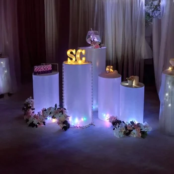 Colonne cylindrique : Arrangement de mariage avec des colonnes cylindriques éclairées par des LED, dégageant une lumière douce et colorée. Les colonnes sont décorées de fleurs artificielles blanches et roses. Sur la colonne centrale, les lettres lumineuses "SC" sont en évidence. D'autres colonnes affichent des éléments décoratifs, tels que des bougies et des petits accessoires dorés, créant une ambiance élégante et romantique.