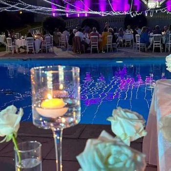 Décoration de mariage en plein air autour d'une piscine, avec des roses blanches et des bougies flottantes dans des photophores en verre. En arrière-plan, des tables rondes élégamment dressées pour les invités, illuminées par des guirlandes lumineuses suspendues. L'éclairage violet crée une ambiance festive et romantique, se reflétant dans l'eau de la piscine.